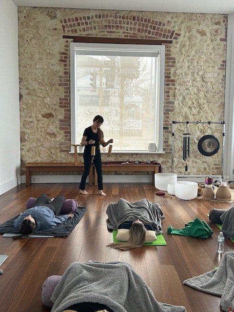 Danielle stands with an instrument while participants rest on mats in a limestone-walled room.
