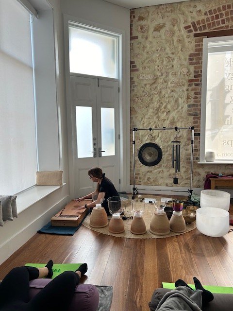 Danielle sits among bowls and chalices beside tall windows as participants listen.