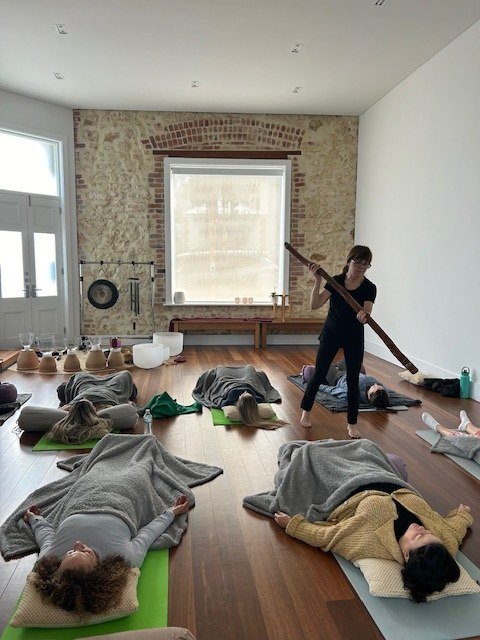 Danielle extends her arms while playing an instrument for a group resting on mats.