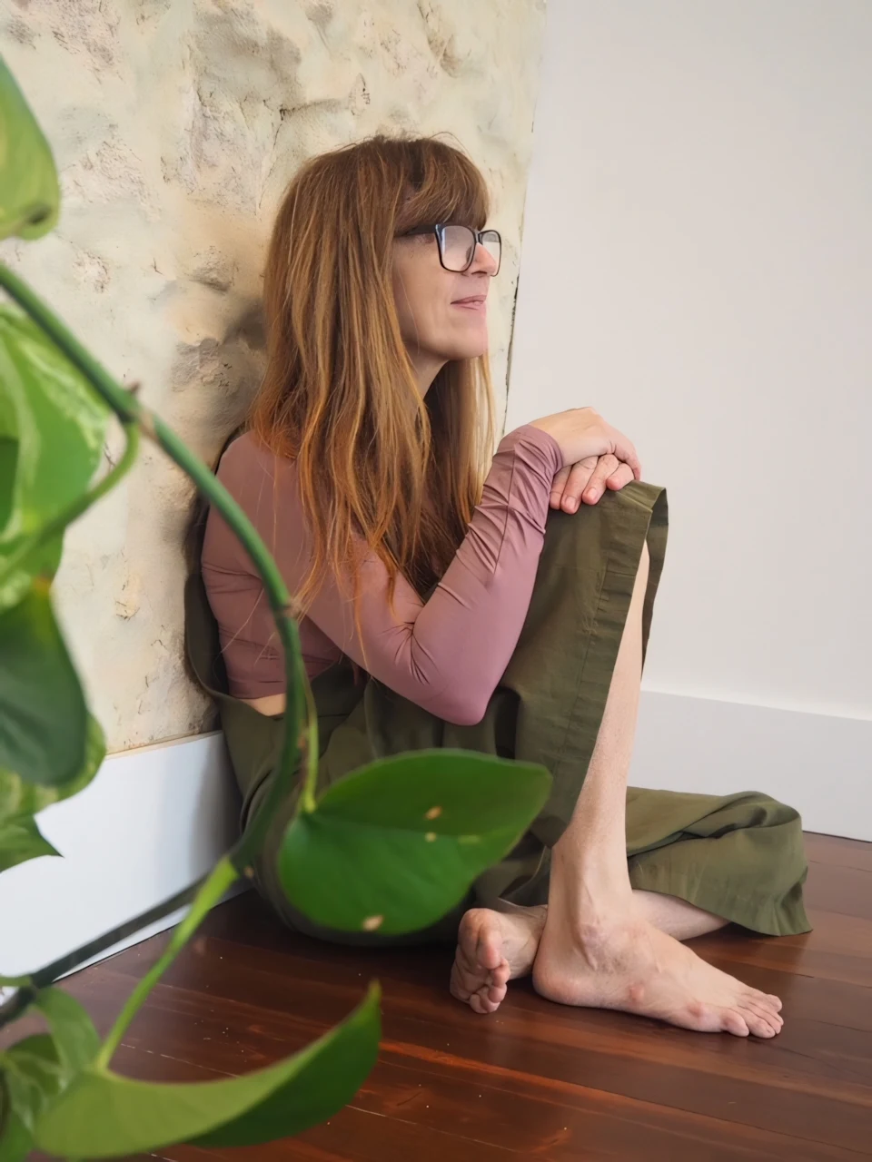 Danielle seated beside a textured wall, framed by green leaves.