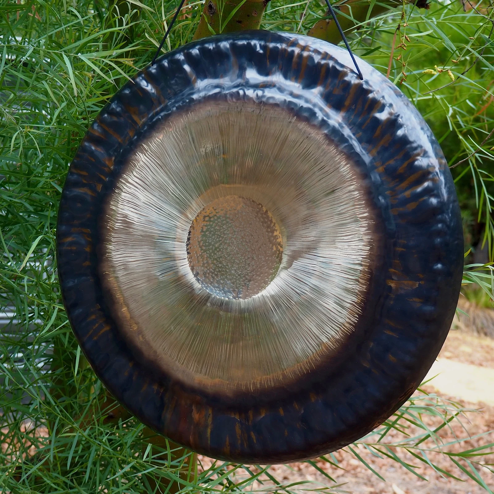 A dark bronze gong suspended among green foliage.