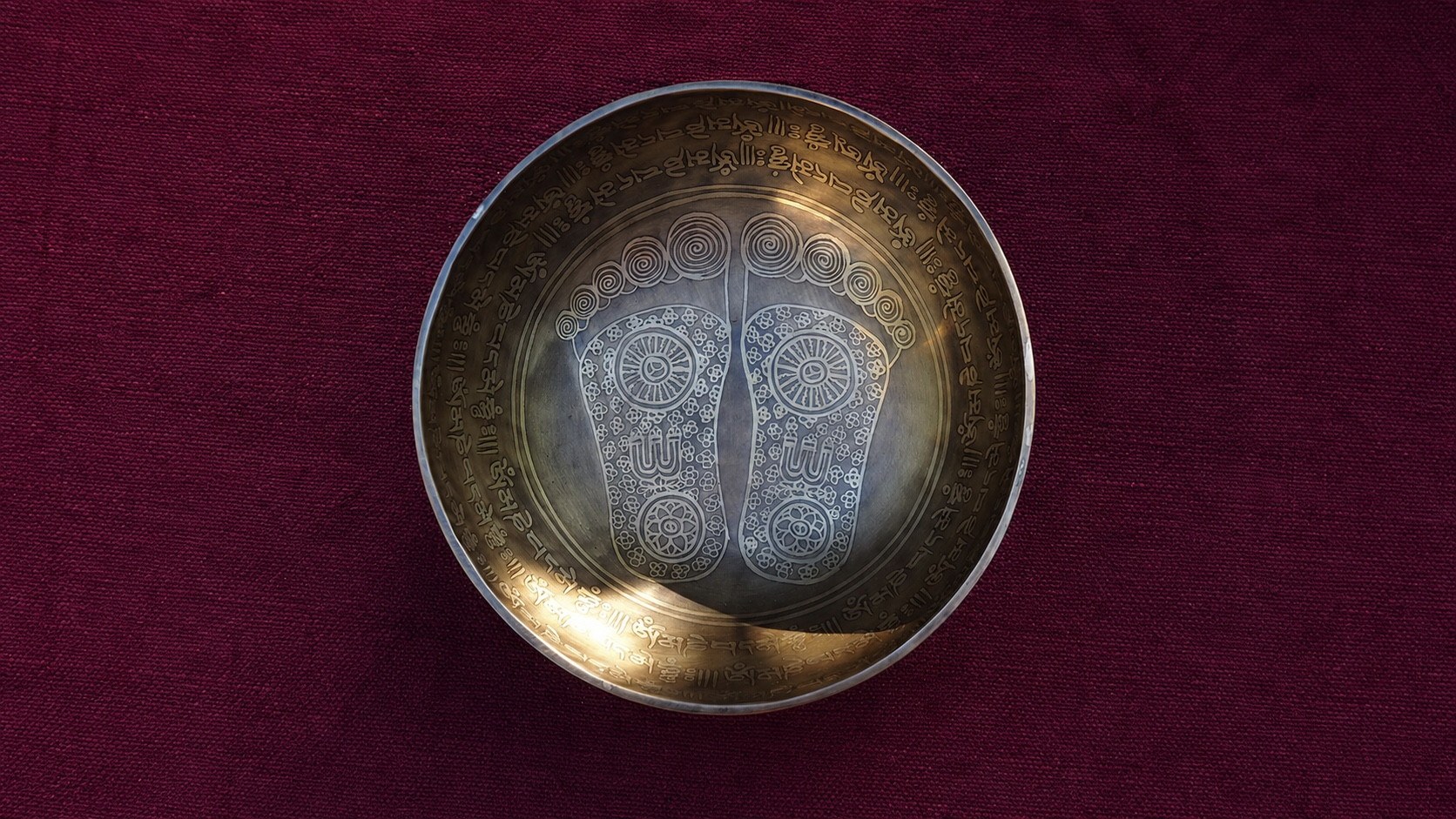 A large engraved singing bowl decorated with two ceremonial footprints on a deep red textile.