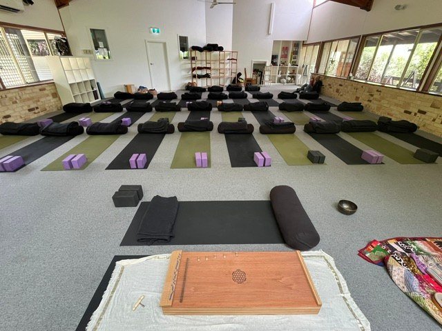 Rows of mats and bolsters face a monochord in a bright yoga studio overlooking trees.
